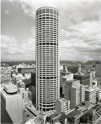 Black and white aerial view of a tall, cylindrical skyscraper amidst city buildings.
