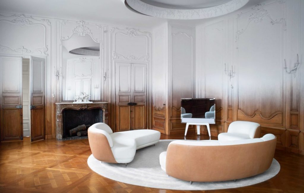 Elegant room blending classical molding with modern design, featuring ombre-painted walls transitioning from dark wood to white, parquet floors, a marble fireplace, and curved modern white furniture, best interior designers in Paris